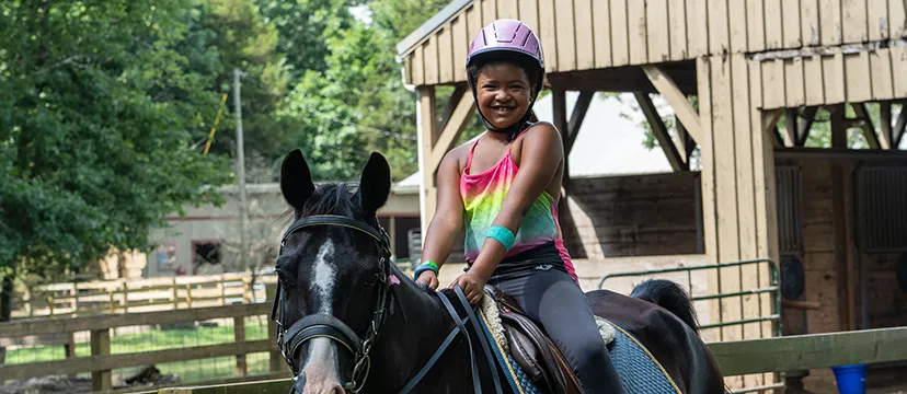 A smiling girl in a pink helmet rides on a black horse at a YMCA ranch camp.